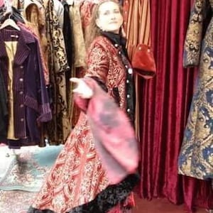 May include: A woman wearing a red and gold patterned coat with black trim. The coat is part of a collection of vintage clothing on display, including coats and jackets in various colors and patterns.