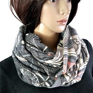 Peut inclure: Une écharpe infinie présentant un motif tissé dans des tons de noir, blanc, gris et rose sourd. Le tissu a un aspect texturé avec des formes et des lignes abstraites. L'écharpe est drapée en plusieurs boucles autour de la zone du cou.