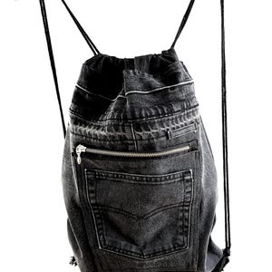 Può includere: Zaino con coulisse in denim nero con tasca con cerniera e tasca posteriore. La borsa è realizzata con jeans di denim riciclati e ha spallacci in corda nera. La borsa è appesa a una gruccia bianca.