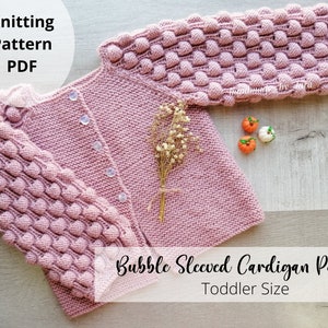 Könnte beinhalten: Ein rosa gestrickter Cardigan mit Ballonärmeln. Der Cardigan hat Knöpfe vorne und ist in Kindergröße. Das Strickmuster ist als PDF-Download erhältlich.