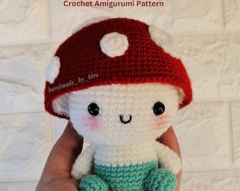 May include: A crocheted amigurumi mushroom toy with a red cap and white spots. The mushroom has a white body and a green skirt. The toy is being held in a hand.