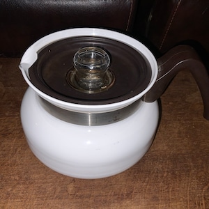 May include: A white glass coffee pot with a dark brown handle and lid. The lid has a clear glass knob in the center. The pot sits on a wooden surface.