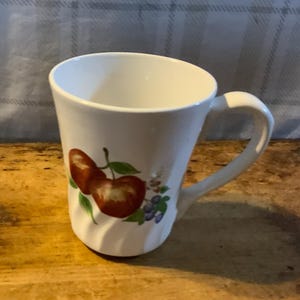 May include: White ceramic mug with a curved handle. The mug features a design of two red apples with green leaves and small purple berries. The mug is set on a wooden surface.