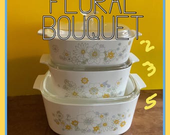 Op de afbeelding: Een set van drie witte ovenschaaltjes met bloemmotieven in tinten geel, blauw en grijs. De schaaltjes zijn op elkaar gestapeld, met het grootste schaaltje onderaan en het kleinste schaaltje bovenaan. De schaaltjes zijn gelabeld met "Floral Bouquet" en hebben de nummers "2", "3" en "5" erop geschreven.