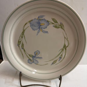 May include: A white ceramic plate with a light blue iris floral design. The plate has a green border and is displayed on a metal stand. The iris flowers have yellow centers and green leaves.