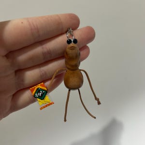 May include: A whimsical wooden keychain shaped like a figure with black eyes and string arms and legs. The figure is holding a small, colorful package with Japanese text. The keychain is attached to a silver ring.
