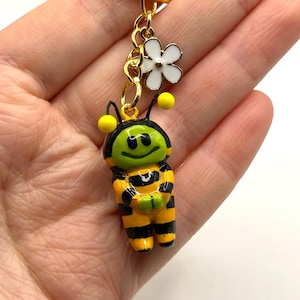 May include: A gold keychain with a small, smiling bee charm. The bee is yellow and black with green eyes and a white flower charm.