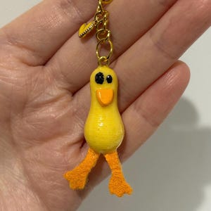 以下が含まれることがあります： 黒い目とオレンジ色のくちばし、オレンジ色の足を持つ黄色いゴム製のあひるのキーホルダー。アヒルは小さなハート型のチャームが付いた金色のチェーンに繋がっています。