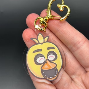 Puede incluir: Un llavero de acrílico transparente con la cabeza de un pájaro amarillo de dibujos animados, con un pico naranja brillante, cuencas oculares oscuras y una boca abierta que muestra los dientes. Incluye un cierre de mosquetón dorado en forma de corazón.