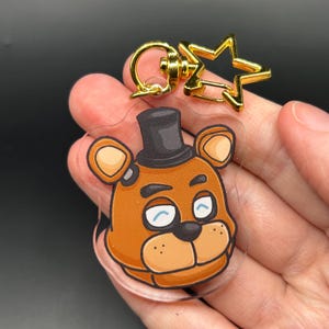 Puede incluir: Un llavero de acrílico transparente con una cabeza de oso de dibujos animados con pelaje marrón, un sombrero de copa negro y una expresión de guiño. El llavero está unido a un cierre de metal dorado con forma de estrella para colgarlo.
