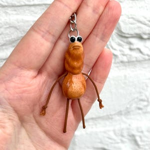 Puede incluir: Un llavero artesanal que presenta una criatura antropomórfica caprichosa. La figura tiene un cuerpo marrón texturizado, ojos negros y brazos y piernas de cuerda marrón. El llavero está unido a un anillo de metal plateado.