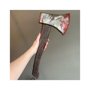 May include: A brown wooden-handled axe with a red, white, and silver painted head. The axe is held by a hand in front of a gray wall.