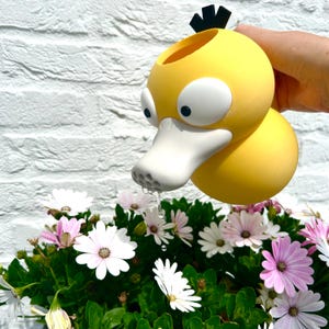Puede incluir: Una regadera amarilla con forma de pato, grandes ojos blancos, un pico blanco con pequeños orificios para el flujo de agua y tres plumas negras en la parte superior. La regadera está inclinada, vertiendo un chorro de agua sobre flores blancas.