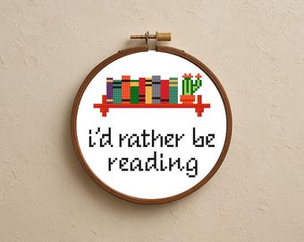 May include: Cross-stitch hoop art with the phrase "I'd rather be reading." The design features a shelf with colorful books and a cactus. The hoop is made of wood and is displayed against a neutral background.