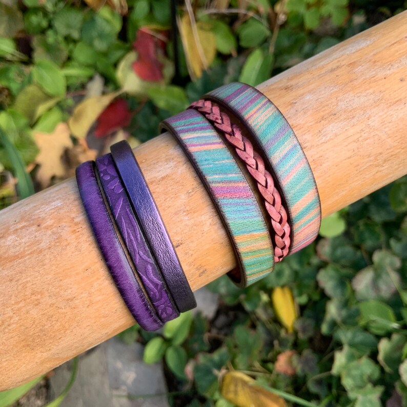 Pu&ograve; includere: Tre braccialetti di pelle impilati su una superficie di legno. Il braccialetto superiore &egrave; un braccialetto di pelle rosa intrecciato. Il braccialetto centrale &egrave; un braccialetto di pelle a strisce multicolori. Il braccialetto inferiore &egrave; un braccialetto di pelle viola scuro con un motivo floreale.