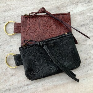May include: Two small leather pouches with zipper closures and decorative embossed floral patterns. The pouch on top is brown with a gold ring attached to the strap. The pouch on the bottom is black with a gold ring attached to the strap.
