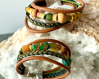 May include: Two stacked leather bracelets with silver clasps. The bracelets feature brown leather, braided green cord, and colorful wooden beads. One bracelet has a mix of yellow, green, and red beads. The other has green and yellow beads.