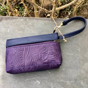 May include: A small purple leather clutch with a navy blue flap and a gold zipper. The clutch has a floral embossed design and a navy blue strap with a gold clasp.