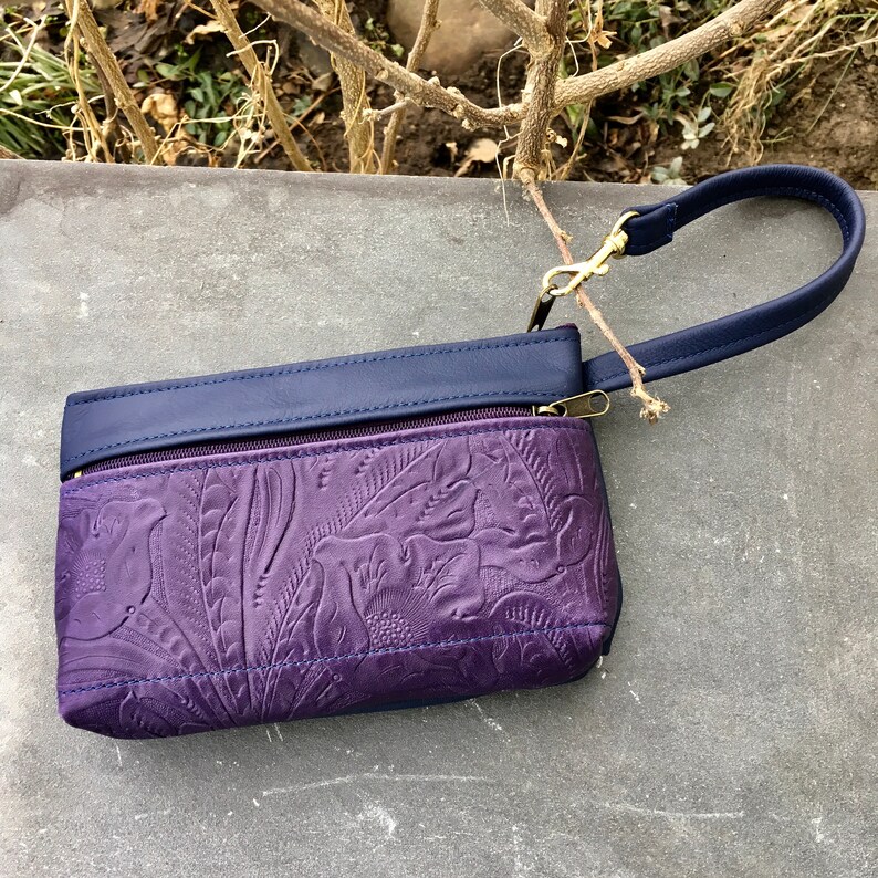 May include: A small purple leather clutch with a navy blue flap and a gold zipper. The clutch has a floral embossed design and a navy blue strap with a gold clasp.