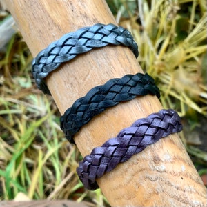 Pode incluir: Três pulseiras de couro trançado em cinza, preto e roxo. As pulseiras são exibidas em um pino de madeira em um fundo de grama verde e folhagem. As pulseiras são um acessório simples, mas elegante.