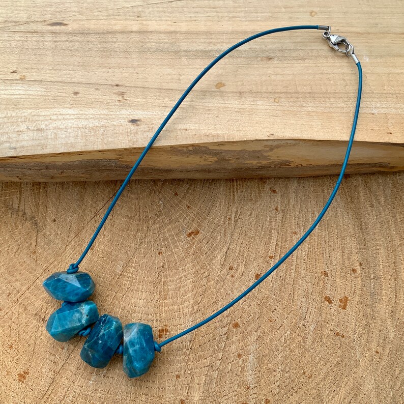 May include: A blue gemstone necklace featuring five faceted, irregular-shaped stones strung on a matching blue cord. The necklace has a silver-colored clasp and is displayed on a wooden surface.