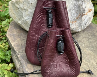 May include: Two burgundy leather pouches with black drawstrings and black gemstone accents. The pouches are embossed with a floral pattern.