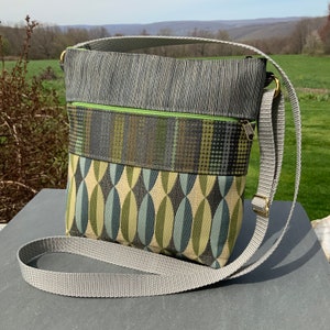 Op de afbeelding: Een grijze en groene crossbody tas met een lange verstelbare band. De tas heeft een ritssluiting en een klep met een geometrisch ontwerp.