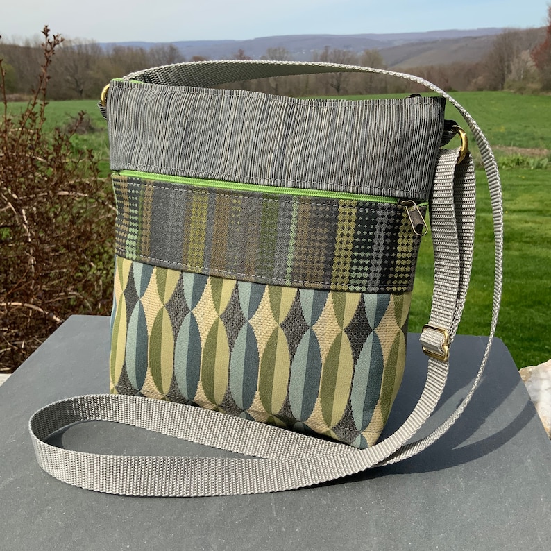 Op de afbeelding: Een grijze en groene crossbody tas met een lange verstelbare band. De tas heeft een ritssluiting en een klep met een geometrisch ontwerp.