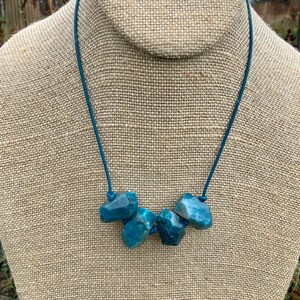 May include: A necklace featuring five rough-cut, teal-colored stones strung on a teal cord. The stones are irregularly shaped and have a slightly textured appearance. The necklace is displayed on a neutral-toned mannequin.