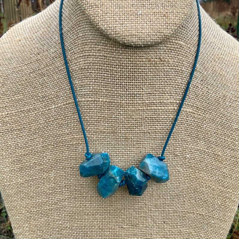 May include: A necklace featuring five rough-cut, teal-colored stones strung on a teal cord. The stones are irregularly shaped and have a slightly textured appearance. The necklace is displayed on a neutral-toned mannequin.