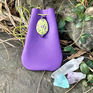 May include: A purple leather pouch with a green and yellow stone pendant. The pouch has a drawstring closure and is sitting on a rock with three crystals: a clear quartz crystal, a light blue crystal, and a purple amethyst crystal.