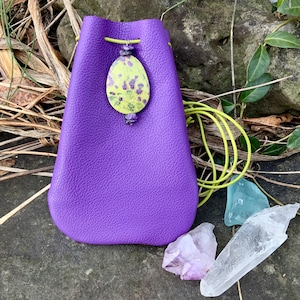 May include: A purple leather pouch with a green drawstring and a yellow and purple decorative bead. The pouch is sitting on a rock with a clear quartz crystal and an amethyst crystal next to it.
