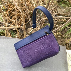 May include: A small purple leather purse with a blue leather flap and a gold zipper. The purse has an embossed floral design and a top handle.