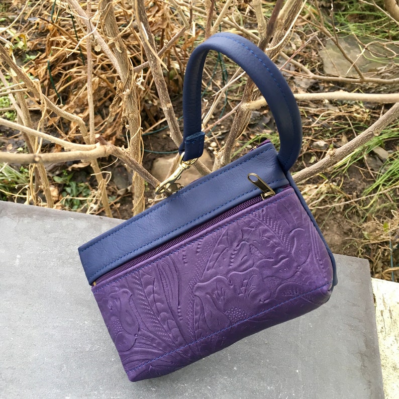 May include: A small purple leather purse with a blue leather flap and a gold zipper. The purse has an embossed floral design and a top handle.