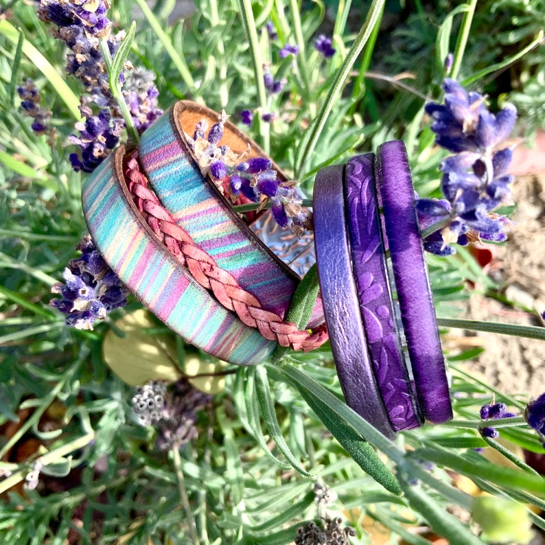 Pu&ograve; includere: Un set di quattro braccialetti in pelle in tonalit&agrave; viola e blu. I braccialetti sono impilati uno sopra l'altro e presentano diverse trame e motivi.