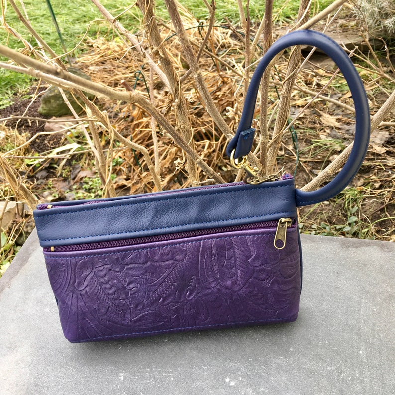 May include: A purple leather clutch with a blue leather top flap and a blue leather top handle. The clutch has a floral embossed design and two zippered compartments.
