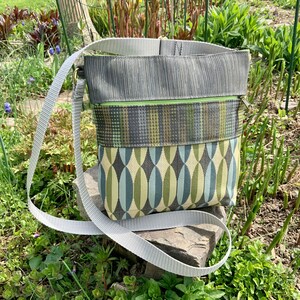 Op de afbeelding: Een grijze en groene crossbody tas met een lange verstelbare schouderband. De tas heeft een ritssluiting en een klep met een drukknopsluiting.