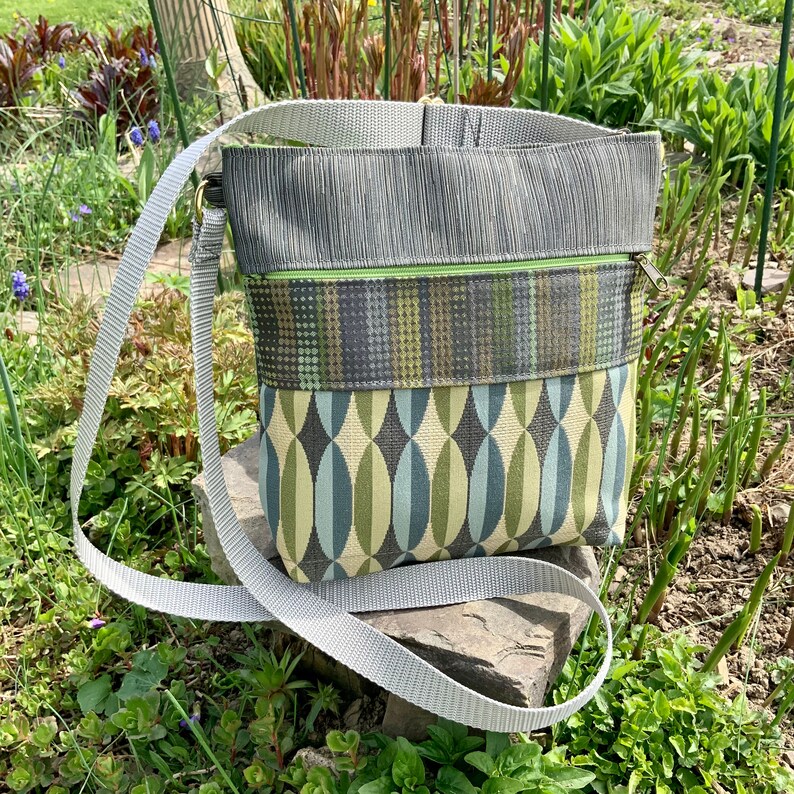 Op de afbeelding: Een grijze en groene crossbody tas met een lange verstelbare schouderband. De tas heeft een ritssluiting en een klep met een drukknopsluiting.