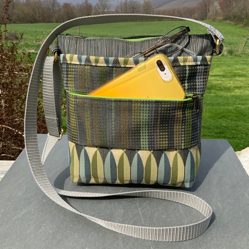 Op de afbeelding: Een grijze en groene crossbody tas met een grijze verstelbare schouderband. De tas heeft een ritsvak en een telefoonvak. Een gele telefoon en een bril bevinden zich in de tas.