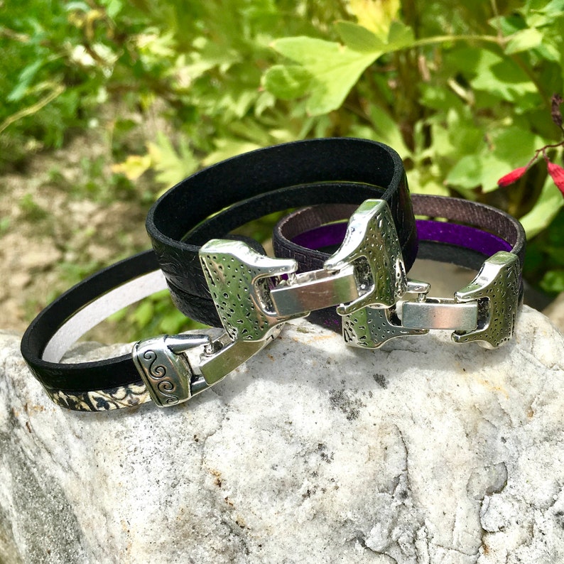 May include: Three leather bracelets with silver clasps. The bracelets are layered on top of each other. The top bracelet is black, the middle bracelet is purple, and the bottom bracelet is black with a floral pattern.