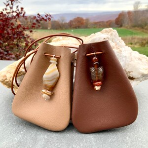 May include: Two small leather pouches, one tan and one brown, with beaded accents. The tan pouch has a diamond-shaped stone pendant with tan and cream stripes. The brown pouch features a turtle-shaped bead. Both have leather drawstring cords.