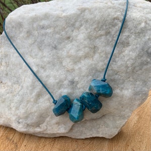 May include: A necklace featuring five faceted, teal-colored gemstone beads strung on a dark blue cord. The gemstones have irregular shapes and are spaced evenly along the cord. The necklace is displayed against a white stone background.