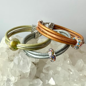 May include: Three leather bracelets with colourful glass beads. The bracelets are in shades of brown, green and silver.