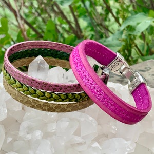 Pode incluir: Um conjunto de quatro pulseiras de couro em tons de rosa e verde. As pulseiras estão empilhadas umas sobre as outras e apresentam diferentes texturas e padrões. As pulseiras são feitas de couro genuíno e têm um fecho prateado.