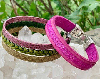 Può includere: Un set di quattro braccialetti in pelle in tonalità rosa e verde. I braccialetti sono impilati uno sopra l'altro e presentano diverse trame e motivi. I braccialetti sono realizzati in vera pelle e hanno una chiusura argentata.