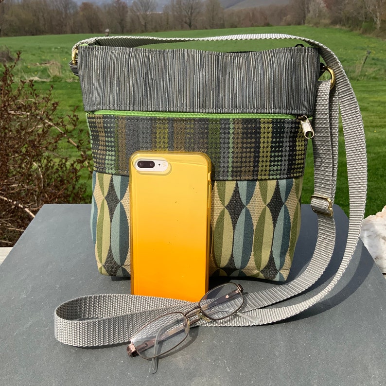 Op de afbeelding: Een grijze en groene schoudertas met een ritsvak en een geometrisch patroon. De tas wordt getoond met een gele telefoon en een bril erin.