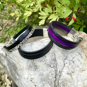 May include: Three leather bracelets with silver clasps. One bracelet is black with a silver embossed design, one is black with a purple stripe, and one is all black.