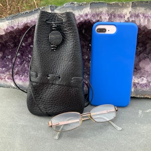 K&ouml;nnte beinhalten: Eine schwarze Ledertasche mit Kordelzug und einer dekorativen Holzperle. Die Tasche liegt auf einem Tisch neben einer blauen Handyh&uuml;lle und einer Brille.