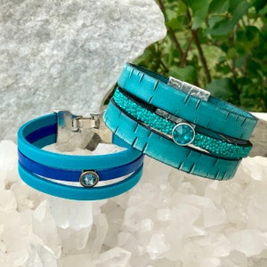 May include: Two leather bracelets with turquoise blue and dark blue bands and silver accents. Each bracelet features a turquoise blue rhinestone.