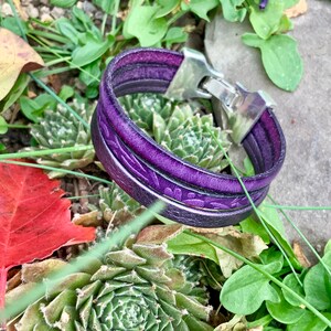 Pu&ograve; includere: Un braccialetto in pelle viola con una chiusura in argento. Il braccialetto &egrave; composto da due fili di pelle e presenta un motivo floreale.